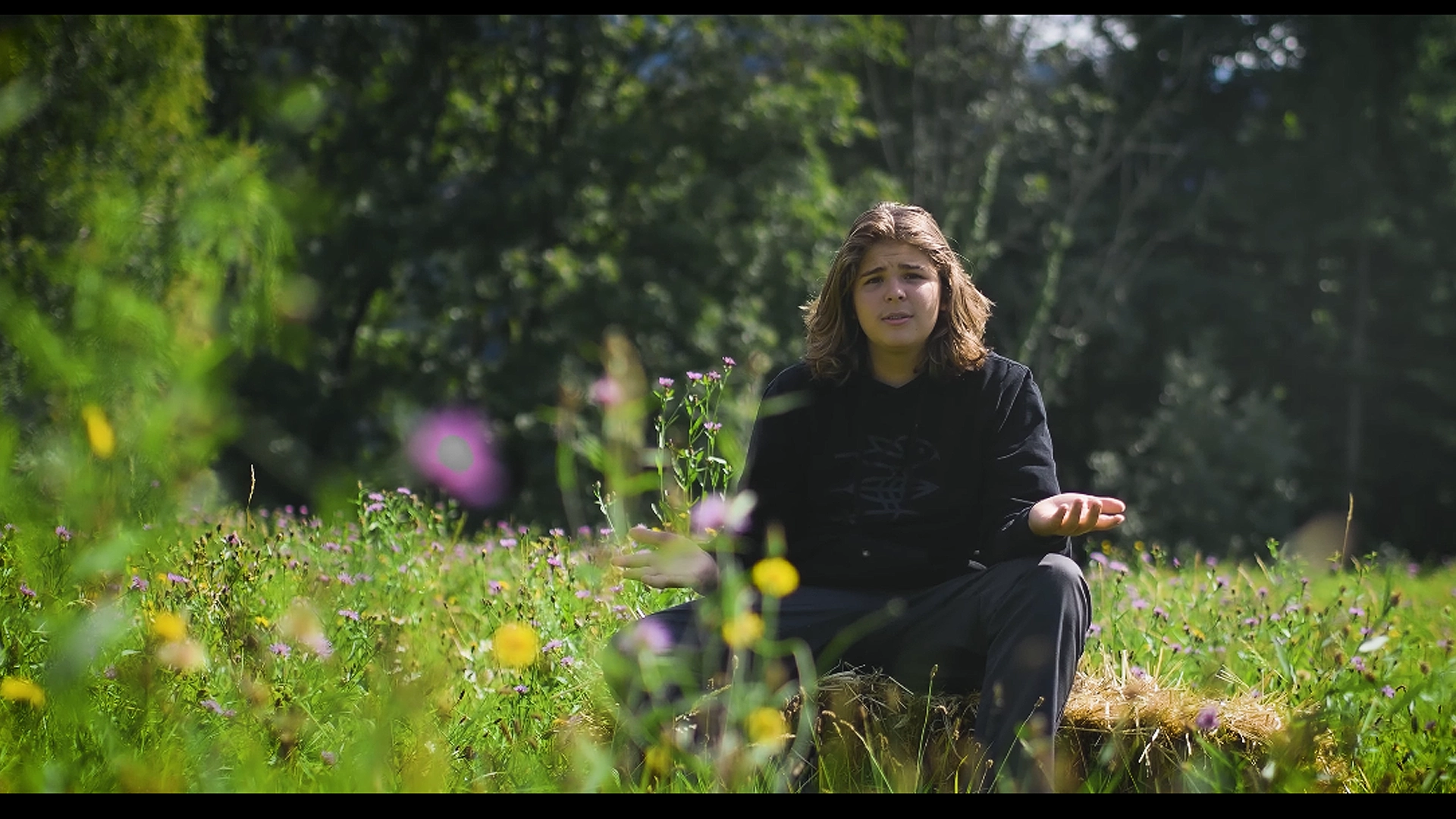 Szenenbild aus dem Kurzfilm 'Artenvielfalt' von Simeo Betschart: Simeo Betschart auf einem Heuballen in einer blühenden Blumenwiese vor Waldkulisse